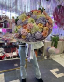 "Large pastel bouquet featuring roses, hydrangeas, and chrysanthemums wrapped in lilac paper with delicate ribbons." "Elegant mixed floral arrangement in soft pink, peach, and lilac tones." "A person holding a voluminous pastel flower bouquet in a floral shop setting."