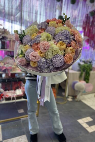 "Large pastel bouquet featuring roses, hydrangeas, and chrysanthemums wrapped in lilac paper with delicate ribbons." "Elegant mixed floral arrangement in soft pink, peach, and lilac tones." "A person holding a voluminous pastel flower bouquet in a floral shop setting."