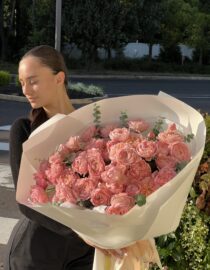 "A young woman with sleek dark hair wearing a black outfit holds a large bouquet of Juliette garden spray roses in soft blush tones, wrapped in luxurious white paper with satin ribbons. The roses have delicate ruffled petals, creating a romantic and timeless floral display."