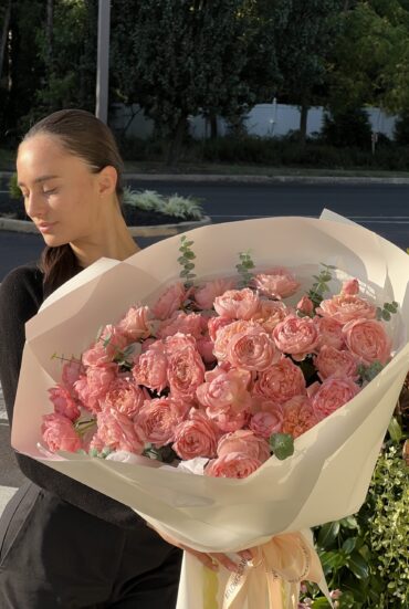 "A young woman with sleek dark hair wearing a black outfit holds a large bouquet of Juliette garden spray roses in soft blush tones, wrapped in luxurious white paper with satin ribbons. The roses have delicate ruffled petals, creating a romantic and timeless floral display."