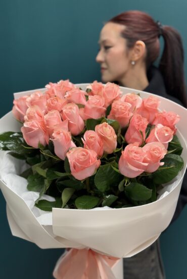 Elegant bouquet of 25 soft pink roses wrapped in white paper with a pastel satin ribbon." "Romantic floral arrangement of fresh pink roses with lush greenery." "A person holding a stunning bouquet of 25 soft pink roses in delicate wrapping."