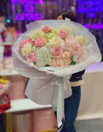 "A luxurious bouquet of soft pink and ivory roses combined with lush white and blush hydrangeas, elegantly wrapped in white designer paper with flowing satin ribbons. The bouquet is held by a person in a stylish outfit, creating a dreamy and sophisticated floral presentation."