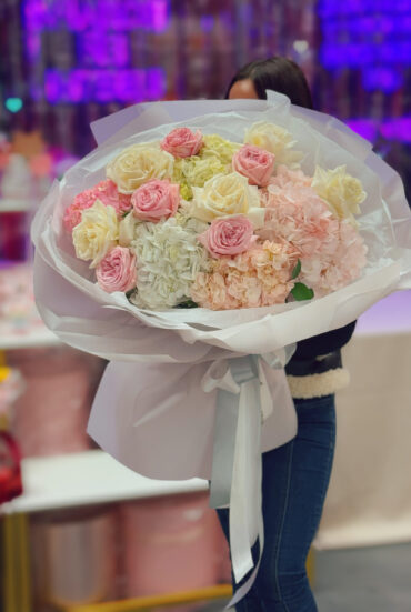 "A luxurious bouquet of soft pink and ivory roses combined with lush white and blush hydrangeas, elegantly wrapped in white designer paper with flowing satin ribbons. The bouquet is held by a person in a stylish outfit, creating a dreamy and sophisticated floral presentation."