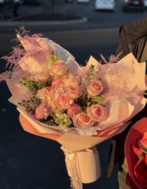 A luxurious bouquet featuring soft peach roses, blush hydrangeas, and pink speckled carnations, accented with airy pink astilbe and delicate greenery. Wrapped in elegant pastel pink designer paper with flowing satin ribbons, the bouquet exudes romance and sophistication, bathed in warm sunlight."