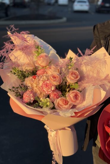 A luxurious bouquet featuring soft peach roses, blush hydrangeas, and pink speckled carnations, accented with airy pink astilbe and delicate greenery. Wrapped in elegant pastel pink designer paper with flowing satin ribbons, the bouquet exudes romance and sophistication, bathed in warm sunlight."