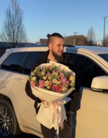 A young man with a stylish beard and a topknot hairstyle holds a large bouquet of lisianthus flowers in shades of pink, lavender, green, and deep magenta. The bouquet is wrapped in crisp white designer paper with soft pink satin ribbons, illuminated by the golden sunset light, creating a warm and elegant ambiance."