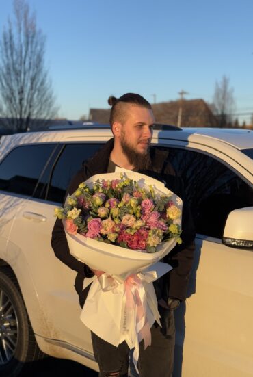 A young man with a stylish beard and a topknot hairstyle holds a large bouquet of lisianthus flowers in shades of pink, lavender, green, and deep magenta. The bouquet is wrapped in crisp white designer paper with soft pink satin ribbons, illuminated by the golden sunset light, creating a warm and elegant ambiance."