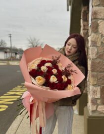 a luxurious bouquet of red and peach roses wrapped in soft pink designer paper with flowing satin ribbons. The bouquet is full and elegantly arranged, perfect for romantic occasions."