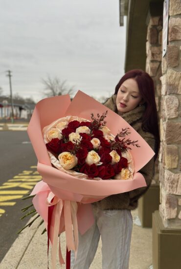 a luxurious bouquet of red and peach roses wrapped in soft pink designer paper with flowing satin ribbons. The bouquet is full and elegantly arranged, perfect for romantic occasions."