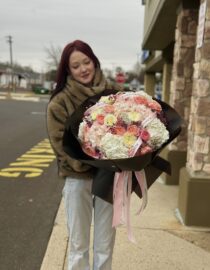 "A luxurious bouquet of soft pink and ivory roses combined with lush white and blush hydrangeas, elegantly wrapped in white designer paper with flowing satin ribbons. The bouquet is held by a person in a stylish outfit, creating a dreamy and sophisticated floral presentation."