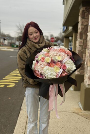 "A luxurious bouquet of soft pink and ivory roses combined with lush white and blush hydrangeas, elegantly wrapped in white designer paper with flowing satin ribbons. The bouquet is held by a person in a stylish outfit, creating a dreamy and sophisticated floral presentation."